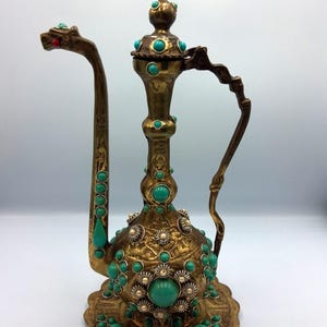 Könnte beinhalten: Verzierter Messingkrug mit türkisfarbenen Akzenten. Der Krug hat einen langen, gebogenen Ausguss mit einem Drachenkopf-Design und einen dekorativen Griff. Der Körper ist mit türkisfarbenen Steinen und floralen Mustern verziert und steht auf einem passenden Sockel.
