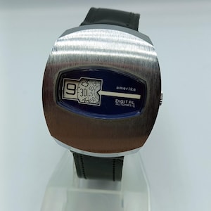 Op de afbeelding: Een vintage digitale horloge met een geborstelde zilverkleurige kast en een zwarte leren band. De wijzerplaat is donkerblauw met witte digitale cijfers en het woord "amerika". Het horloge geeft de tijd 9:30 aan. De woorden "DIGITAL AUTOMATIC" zijn ook zichtbaar.