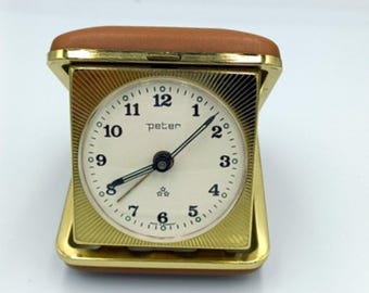 May include: A gold-colored, square-shaped alarm clock with a white face and black hands. The clock is in a brown leather case. The clock face has the word "peter" printed on it.