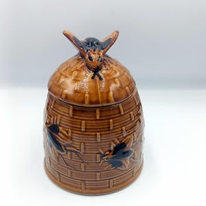 Puede incluir: Un tarro de cerámica marrón con tapa, con un diseño de cesta tejida. La tapa está rematada con un asa en forma de abeja. Ilustraciones de abejas negras están pintadas en el exterior del tarro. Es un artículo de cocina decorativo.