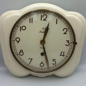 May include: A cream-colored wall clock with a scalloped frame and a circular face. The clock features black Arabic numerals, a brass-toned bezel, and dark metal hands. The face displays the text Junghans and EXA, with a small logo above.