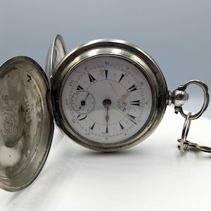 May include: An open, antique silver pocket watch with a white face and black Roman numerals. The watch has a small subdial and a key attached. The watch's cover is open, revealing the inner workings and markings.