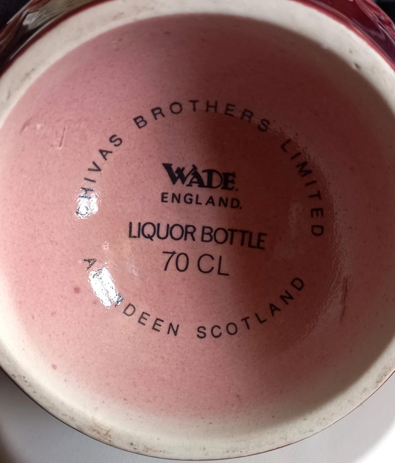 Może przedstawiać: Zbliżenie dna butelki po alkoholu Wade England. Butelka jest w kolorze jasnor&oacute;żowym i ma czarny napis "Chivas Brothers Limited" oraz "Liquor Bottle 70 CL". Widoczny jest r&oacute;wnież napis "Aberdeen Scotland".