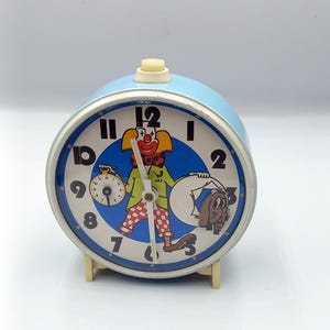 May include: A vintage blue alarm clock with a cartoon clown and dog design on the face. The clock has a white dial with black numbers and hands. The clown is wearing a red bow tie and checkered pants. The clock is approximately 5 inches tall.