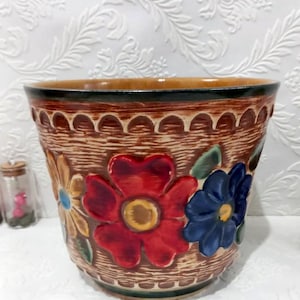 Puede incluir: Una maceta de cerámica marrón con un diseño en relieve de flores en rojo, amarillo, azul y verde. La maceta tiene un borde marrón y verde.