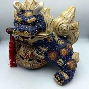 May include: A ceramic Foo Dog figurine, featuring a blue body with gold and red accents. The figurine has a white mane and tail, and a red tassel hangs from its mouth. The Foo Dog is a symbol of protection and prosperity.