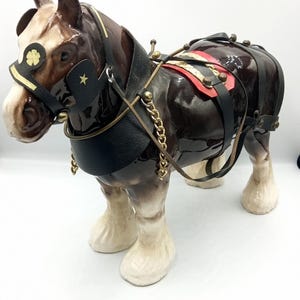 Puede incluir: Figura de cerámica de un caballo Clydesdale, predominantemente marrón con marcas blancas en la cara y las patas. Está adornado con un arnés negro y dorado, una silla de montar roja y detalles de cadena dorada. El caballo está de pie.