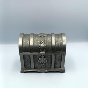 May include: A silver metal treasure chest with a wood grain design. The chest has a hinged lid with a decorative eagle and a lock.