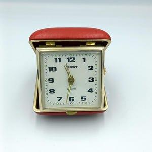 May include: A red, leather-covered, square-shaped alarm clock with a white face and black numbers. The clock face reads "Orient" and "Alarm".