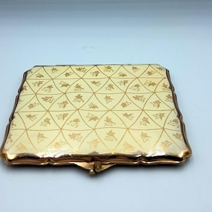 May include: A vintage compact mirror with a cream-colored surface and a gold geometric pattern. The compact has a gold-colored metal frame and clasp. The design features a repeating pattern of gold leaves.