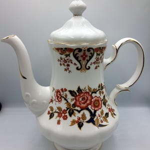 May include: A white porcelain coffee pot with a lid and handle, decorated with floral patterns in red, orange, and navy blue. The pot features gold trim and a decorative finial, showcasing a classic design.