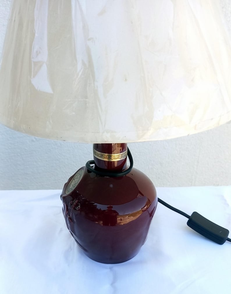 Może przedstawiać: Bordowa ceramiczna lampa stołowa z kremowym abażurem. Podstawa lampy ma teksturowany wz&oacute;r i złoty pasek u g&oacute;ry. Widoczny czarny przew&oacute;d i włącznik/wyłącznik.