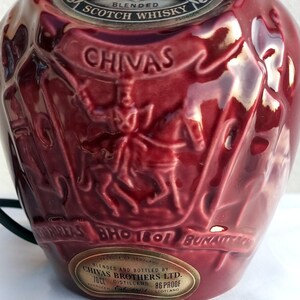Może przedstawiać: Bordowa ceramiczna butelka z wytłoczonymi detalami. Butelka zawiera napis "CHIVAS" w banerze oraz wizerunek osoby na koniu. Butelka posiada r&oacute;wnież metalową etykietę z tekstem. Butelka jest przeznaczona do szkockiej whisky.