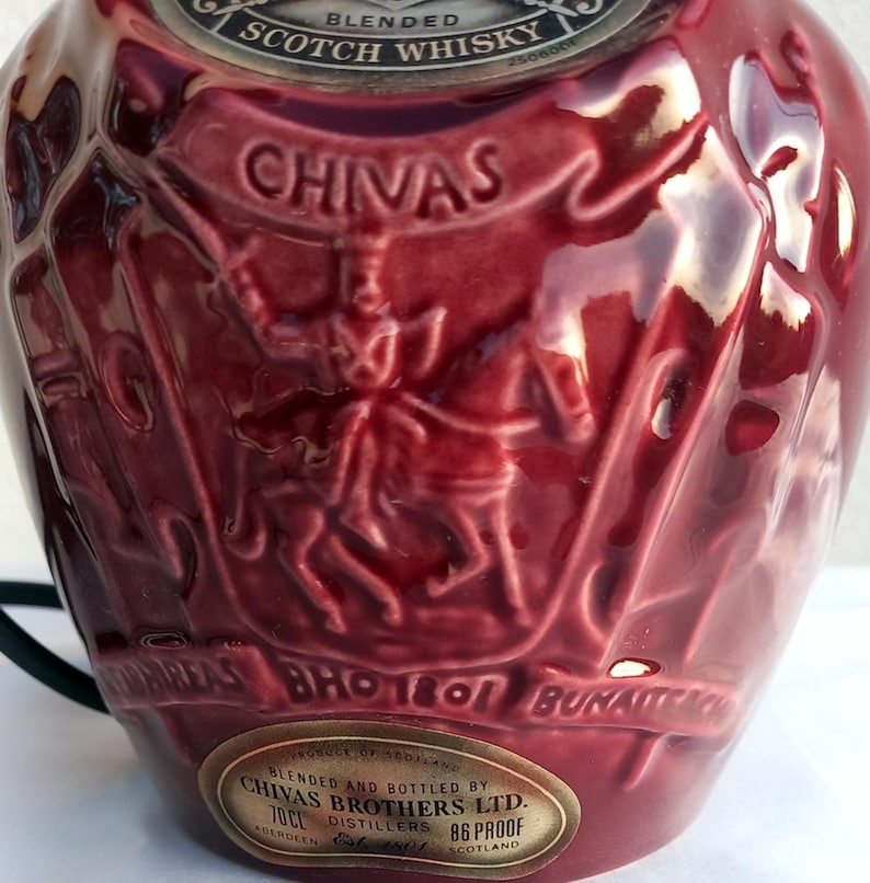 Może przedstawiać: Bordowa ceramiczna butelka z wytłoczonymi detalami. Butelka zawiera napis "CHIVAS" w banerze oraz wizerunek osoby na koniu. Butelka posiada r&oacute;wnież metalową etykietę z tekstem. Butelka jest przeznaczona do szkockiej whisky.