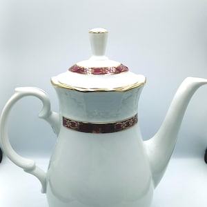 Puede incluir: Cafetera de porcelana blanca con ribete burdeos y dorado. La cafetera tiene un cuerpo redondeado y un pico en el lado derecho. La tapa tiene un pomo en la parte superior.