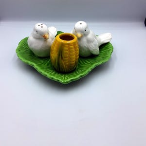 Puede incluir: Un juego de sal y pimienta de cerámica con dos saleros blancos con forma de pájaro, un recipiente amarillo con forma de maíz y una base verde con forma de hoja. El juego está diseñado para uso en mesa.
