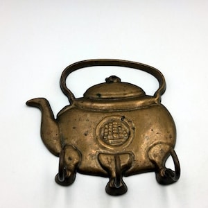 Op de afbeelding: Een vintage bronzen metalen theepot wanddecoratie. De theepot heeft een handvat, een tuit en een decoratief embleem aan de voorkant. Het heeft drie kleine voetjes en een deksel. Het ontwerp suggereert een antiek of decoratief item.