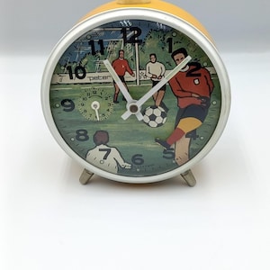 Puede incluir: Reloj despertador amarillo y plateado con un diseño de fútbol. La esfera del reloj presenta una ilustración de un partido de fútbol con jugadores y un balón. El reloj tiene un marco y patas plateadas. La palabra "peter" es visible.