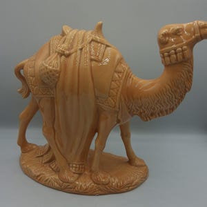 Può includere: Una statuetta di cammello in ceramica color marrone chiaro lucido su una base ovale. Il cammello presenta una sella dettagliata con motivi strutturati, borse pendenti e una briglia. L'intero pezzo ha una finitura liscia e riflettente.