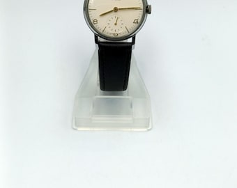 May include: A vintage wristwatch with a white face and black leather strap. The watch face has black Roman numeral hour markers and gold hands. The watch is on a clear plastic stand.