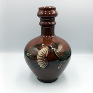 May include: A brown ceramic vase with a bulbous body and a narrow neck. The vase is decorated with a white and tan flower design with green leaves. The vase is approximately 8 inches tall.