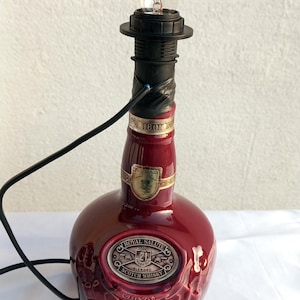 Może przedstawiać: Lampa wykonana z butelki whisky Royal Salute w kolorze bordowym. Lampa posiada czarny uchwyt na żar&oacute;wkę, żar&oacute;wkę i czarny przew&oacute;d zasilający. Butelka ma wytłoczone detale i złote akcenty.