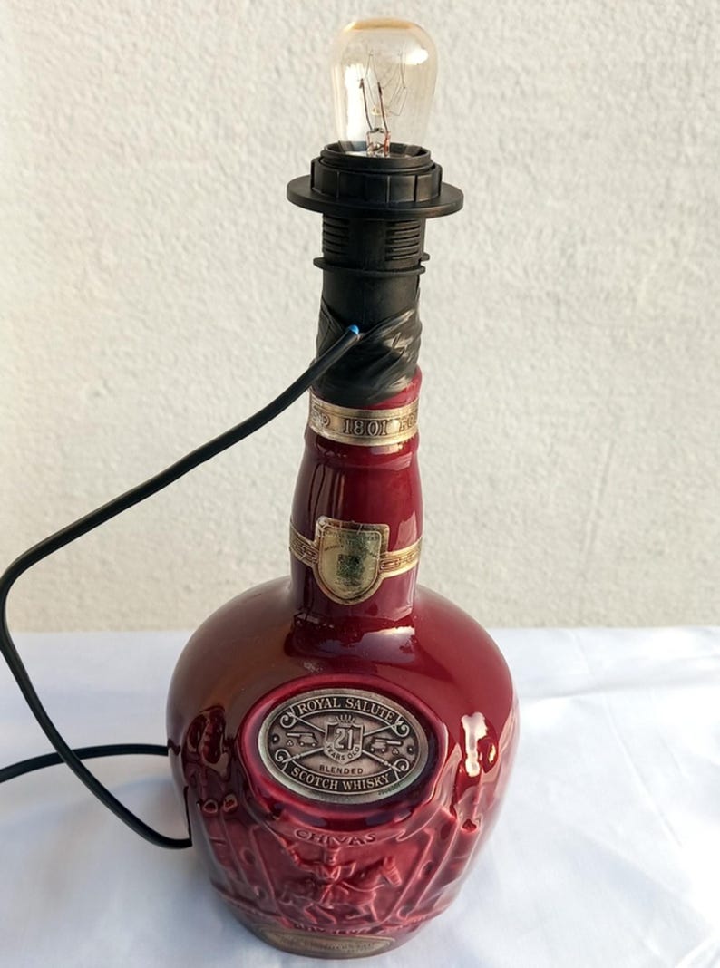 Może przedstawiać: Lampa wykonana z butelki whisky Royal Salute w kolorze bordowym. Lampa posiada czarny uchwyt na żar&oacute;wkę, żar&oacute;wkę i czarny przew&oacute;d zasilający. Butelka ma wytłoczone detale i złote akcenty.