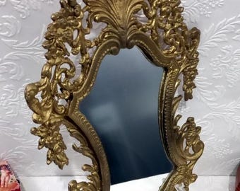 May include: A gold-toned ornate mirror with a decorative frame. The frame is intricately carved with floral and scroll designs.