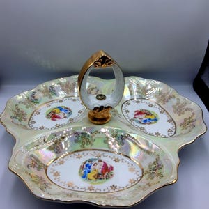 Könnte beinhalten: Ein dreiteiliges Serviertablett mit goldenem Griff. Das Tablett hat eine schillernde Oberfläche mit goldenem Rand und ovale Abschnitte mit farbenfrohen Illustrationen im klassischen Stil. Der Griff ist goldfarben mit einem klaren, ovalen Mittelteil.