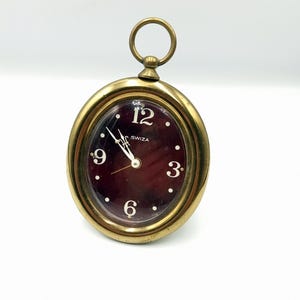 Può includere: Orologio ovale in ottone con quadrante bordeaux. L'orologio ha numeri e lancette bianche, e la parola "SWIZA" è stampata sul quadrante. Un piccolo anello è attaccato alla parte superiore dell'orologio.