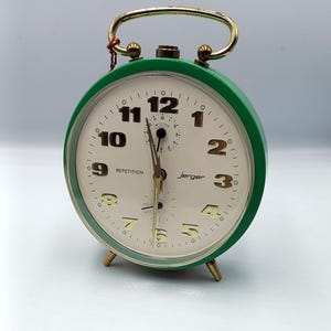 May include: A vintage Jerger alarm clock with a green metal casing and a gold-colored handle. The clock face is cream-colored with black numerals and hands. The word "REPETITION" is printed on the face.
