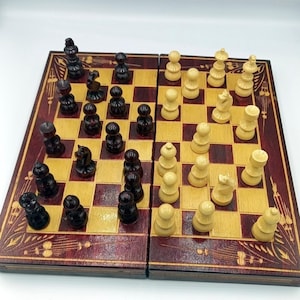 May include: A wooden chessboard with a red and brown finish. The board is open and shows a game in progress with black and white chess pieces.