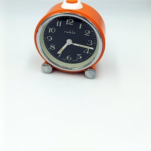May include: Orange Ruhla alarm clock with a black face and silver accents. The clock has a white button on top and silver feet. The clock face displays white numerals and hands. The clock is a retro style.