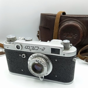 Puede incluir: Una cámara de telémetro FED-2 plateada y negra con una funda de cuero. La cámara tiene un objetivo con una distancia focal de 35 mm y una apertura máxima de f/3,5. La cámara está en buen estado y parece estar bien mantenida.