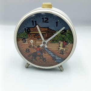 Puede incluir: Un despertador blanco vintage con una esfera temática del oeste. La esfera del reloj presenta una escena pintada de vaqueros, caballos y un edificio. El reloj tiene un botón superior color crema y patas plateadas. El reloj mide aproximadamente 10 cm de alto.