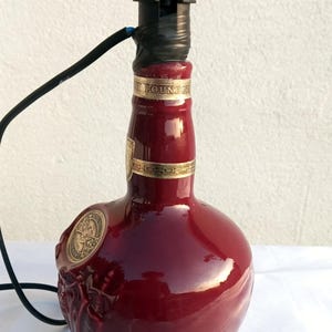 Może przedstawiać: Lampa wykonana z butelki po alkoholu w kolorze bordowym. Butelka ma złote akcenty i ozdobny emblemat. Czarna oprawa lampy i przew&oacute;d są przymocowane do g&oacute;ry. Unikalny element wystroju wnętrz.