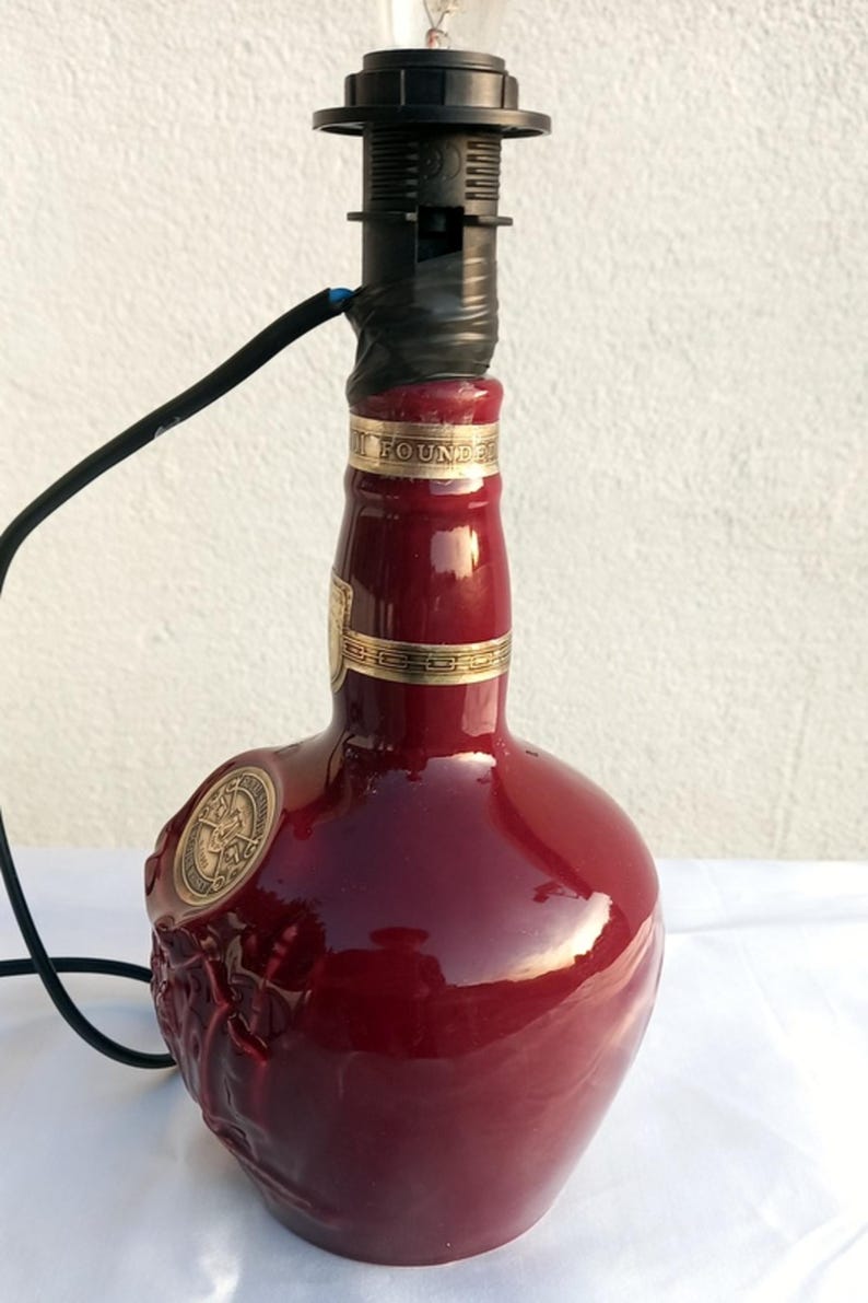 Może przedstawiać: Lampa wykonana z butelki po alkoholu w kolorze bordowym. Butelka ma złote akcenty i ozdobny emblemat. Czarna oprawa lampy i przew&oacute;d są przymocowane do g&oacute;ry. Unikalny element wystroju wnętrz.