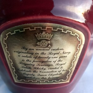 Może przedstawiać: Bordowa butelka ze złotą etykietą. Etykieta zawiera tekst i ilustrację korony. Tekst opisuje whisky Royal Salute, stworzoną dla uczczenia koronacji kr&oacute;lowej Elżbiety.