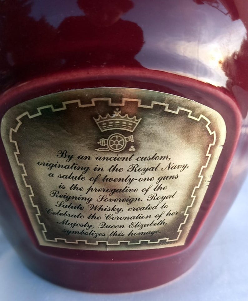 Może przedstawiać: Bordowa butelka ze złotą etykietą. Etykieta zawiera tekst i ilustrację korony. Tekst opisuje whisky Royal Salute, stworzoną dla uczczenia koronacji kr&oacute;lowej Elżbiety.
