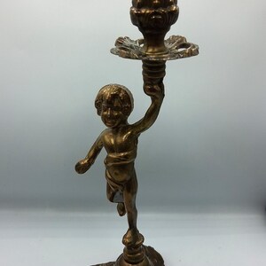 Puede incluir: Un candelabro de metal en tono bronce que presenta la figura esculpida de un querubín sosteniendo una copa para vela en alto. La base es ancha y ornamentada con detalles en relieve floral, sosteniendo la figura central y el soporte.