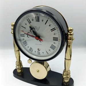 May include: A black and gold desk clock with a round face featuring Roman numerals and a small bird emblem. The clock has gold-colored legs and a pendulum, and sits on a black base. The clock face has black hands and a red second hand.