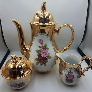 Può includere: Servizio da tè dorato composto da una teiera con coperchio, una zuccheriera con coperchio e una lattiera. Ogni pezzo presenta un motivo floreale di rose rosa e rosse su sfondo bianco.
