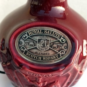 Może przedstawiać: Bordowa butelka Royal Salute 21 Years Old Blended Scotch Whisky. Etykieta zawiera napisy "Royal Salute" i "21 Years Old Blended Scotch Whisky". Butelka ma teksturowaną powierzchnię i dekoracyjny wz&oacute;r.