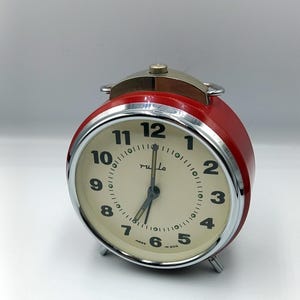 Può includere: Una sveglia rossa vintage con quadrante color crema e numeri neri. L'orologio ha una lunetta cromata e una parte superiore argentata. Il quadrante dell'orologio indica "rula" e "Made in GDR".