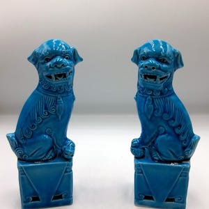 Może przedstawiać: Dwie turkusowe ceramiczne posągi Foo Dog. Każdy pies siedzi na kwadratowym cokole z trójkątnym wzorem. Psy mają szczegółowe cechy, w tym otwarte pyszczki, i są w żywym odcieniu niebieskiego.