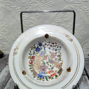 May include: White ceramic decorative plate with a gold rim and a central design featuring two peacocks amidst colorful flowers. The plate is supported by a metal stand, showcasing a vintage aesthetic.