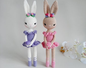 Puede incluir: Dos muñecas de conejo de crochet vestidas de bailarinas. La conejita de la izquierda es blanca con un tutú morado y la conejita de la derecha es marrón con un tutú rosa. Ambas conejitas llevan coronas de flores y sostienen una pequeña rosa.