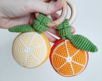 May include: Two crochet baby toys resembling orange and lemon slices, each with a green leaf and attached to a wooden ring. The orange slice is bright orange with white details, and the lemon slice is yellow with white details.