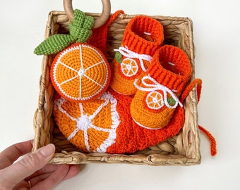 May include: A crochet baby gift set in a wicker basket. The set features orange baby booties, a teething ring with an orange slice, and a matching orange slice. The set is made with orange, white, and green yarn.