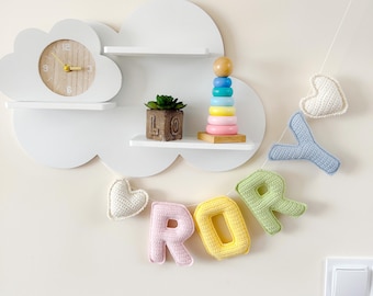 May include: A white cloud-shaped shelf unit with a clock, a small plant, and a wooden stacking toy. A crochet banner spells "ROR" in pastel colors, with a heart and a blue "Y" on either side. The wall is a light beige.
